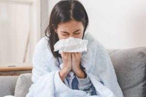 Woman with brown hair snuggled in blankets on the couch blowing her nose into a tissue